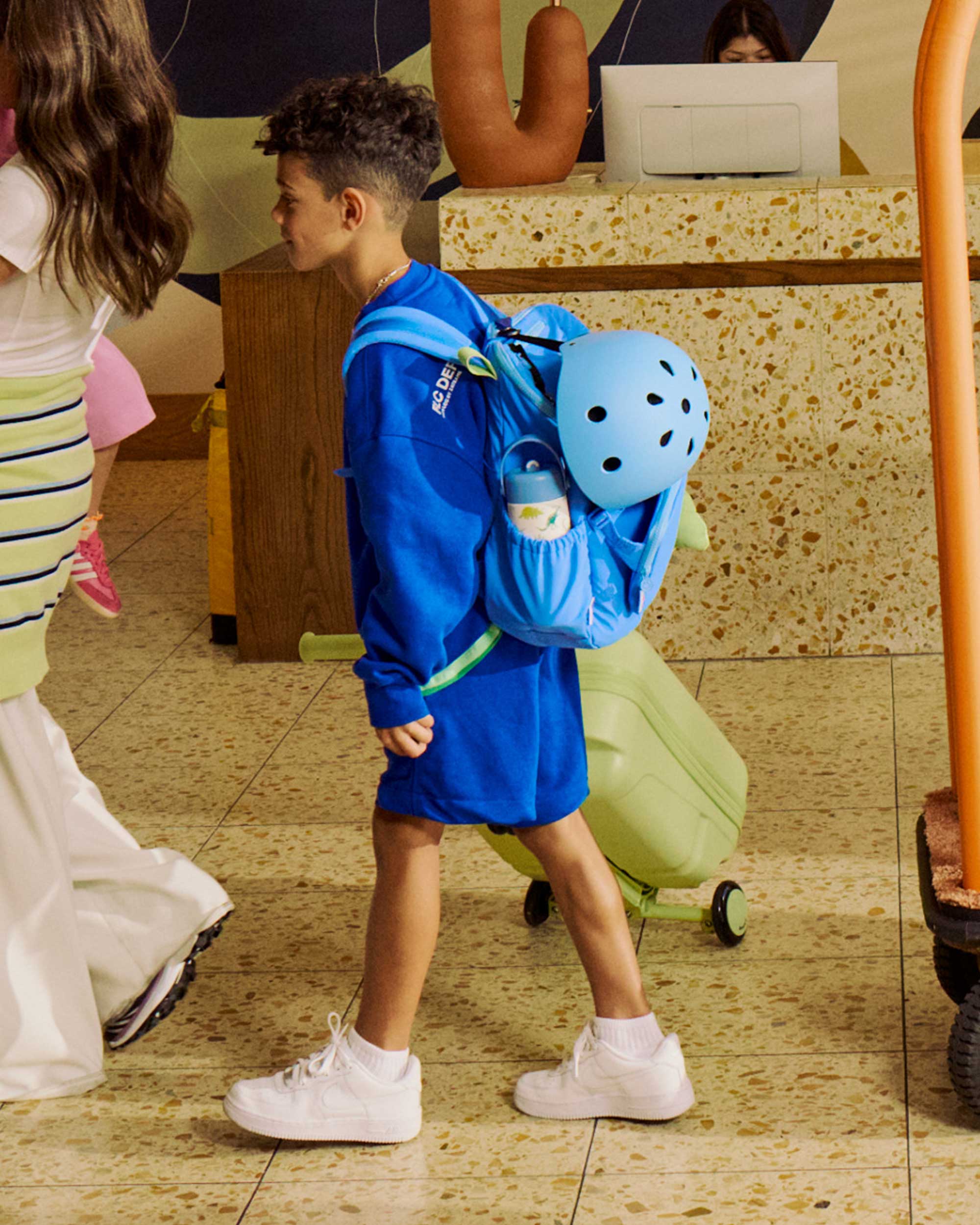 Child in blue outfit with sky blue helmet and backpack walking in a room with tiled floor and furniture.