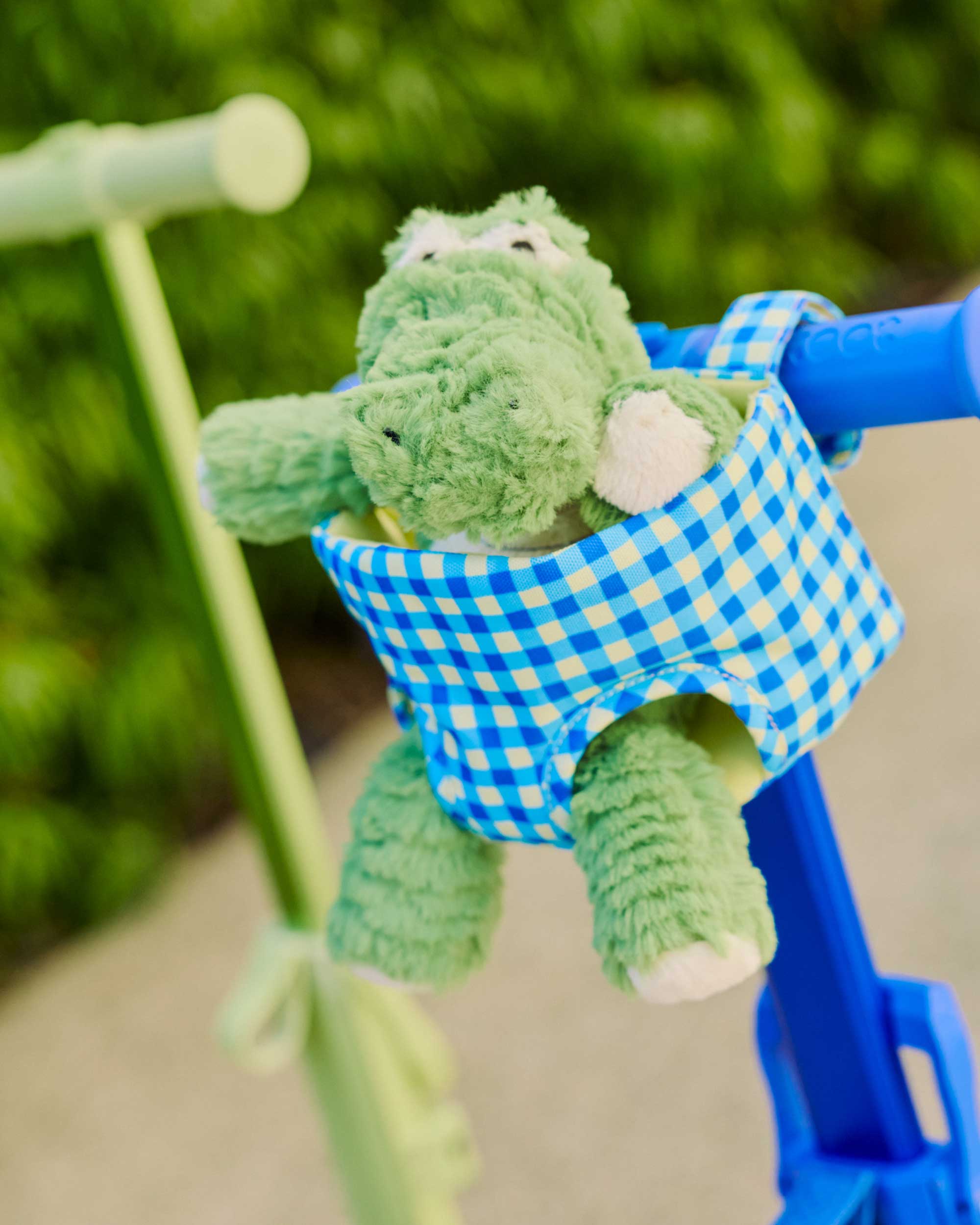 Green plush toy with a blue/green gingham doll carrier attached to a blue skootcase outdoors