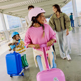 Two children with suitcases at a train station, one wearing a puff pink helmet.