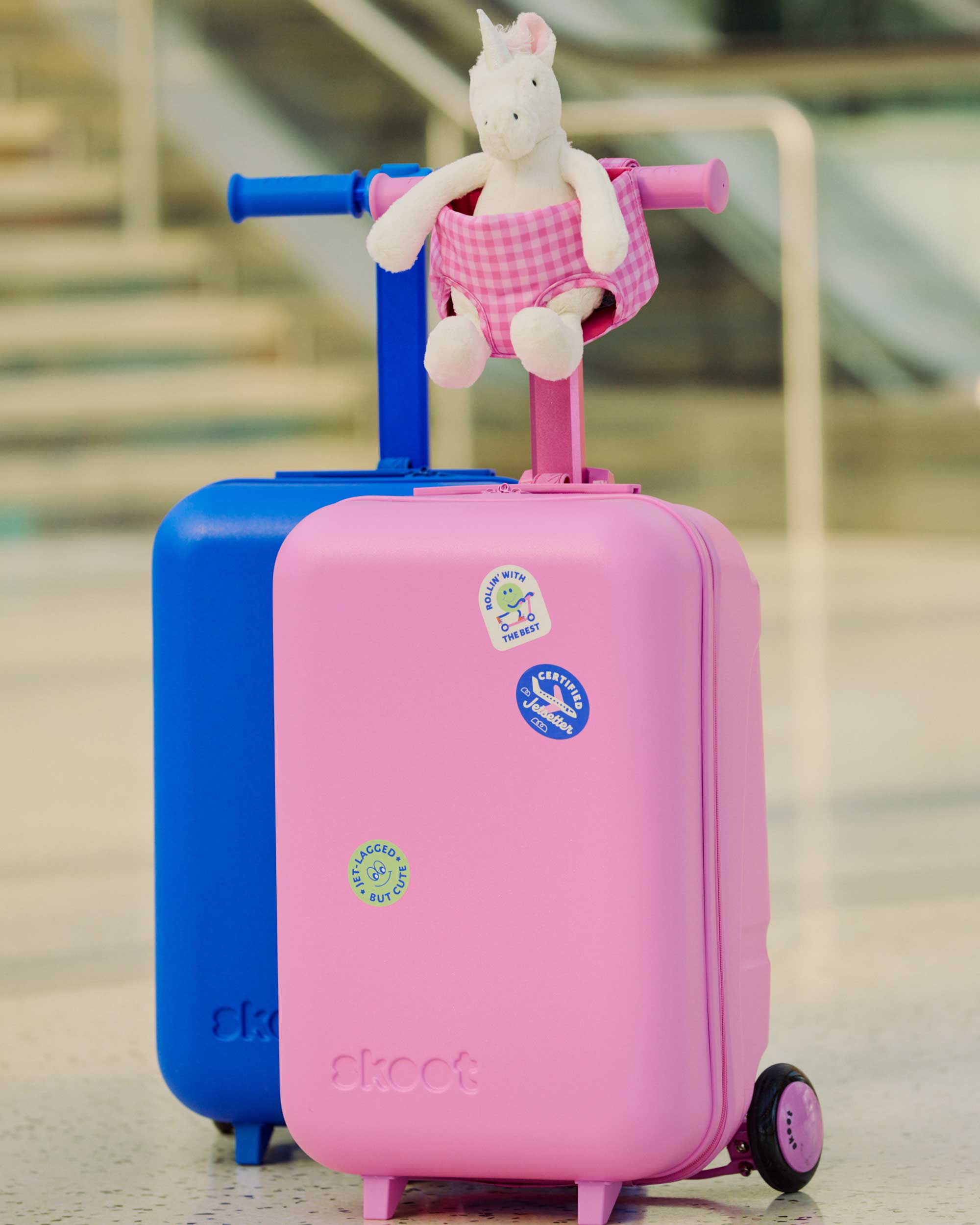 Pink and blue children's skootcases with a unicorn toy on top inside the pink gingham skoot doll carrier, set against an airport background.